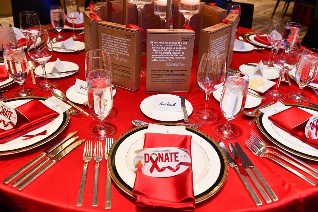 Image of an elegant dining table setting with a red silk tablecloth, gold utensils, a red napkin on white plate with a Donation card, and HIV-self test kits on the table