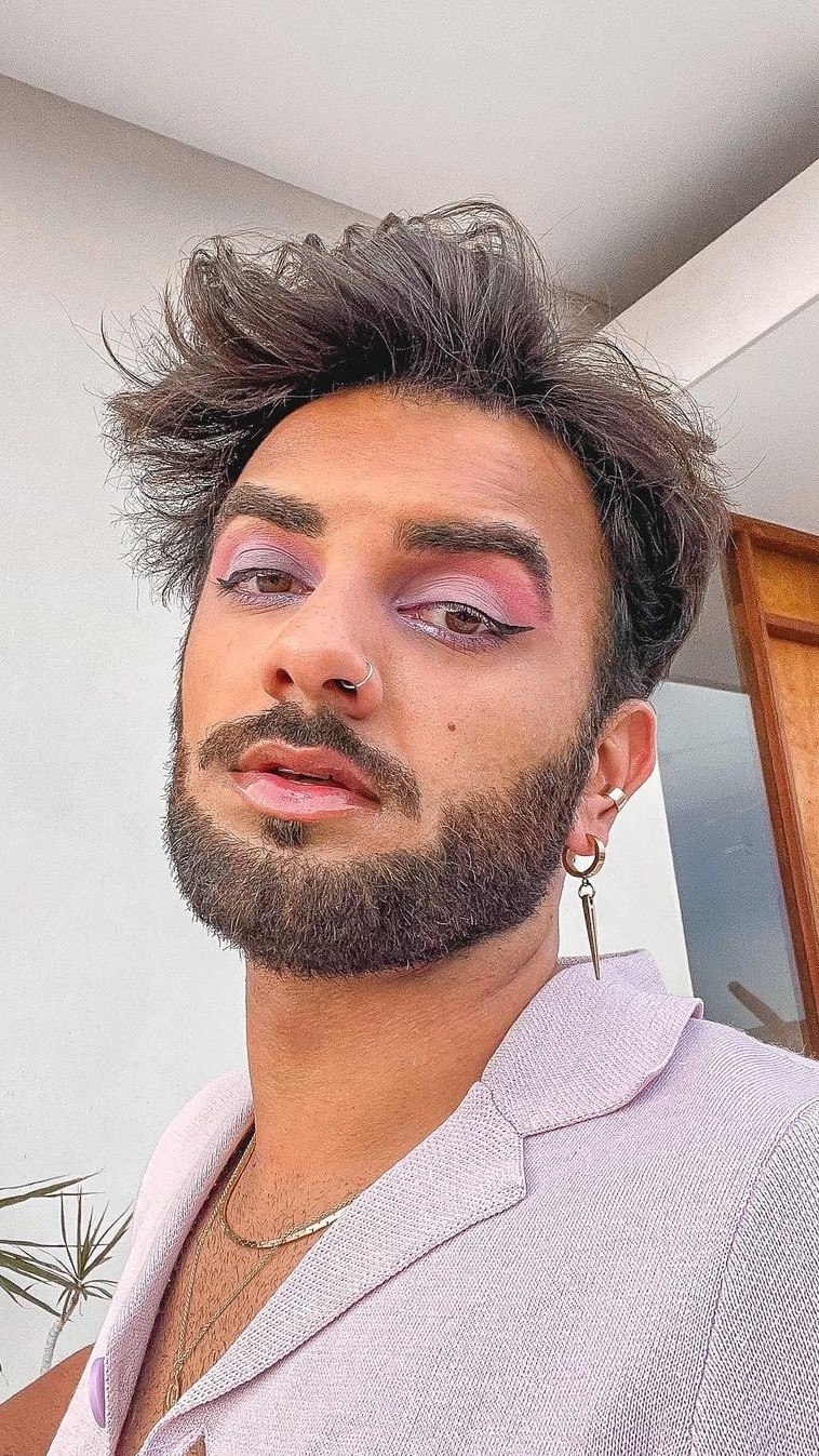 Image of non-binary person of South Asian descent, with dark brown hair wisping off the top of their head. Beard and pinkish-purple eyeshadow.