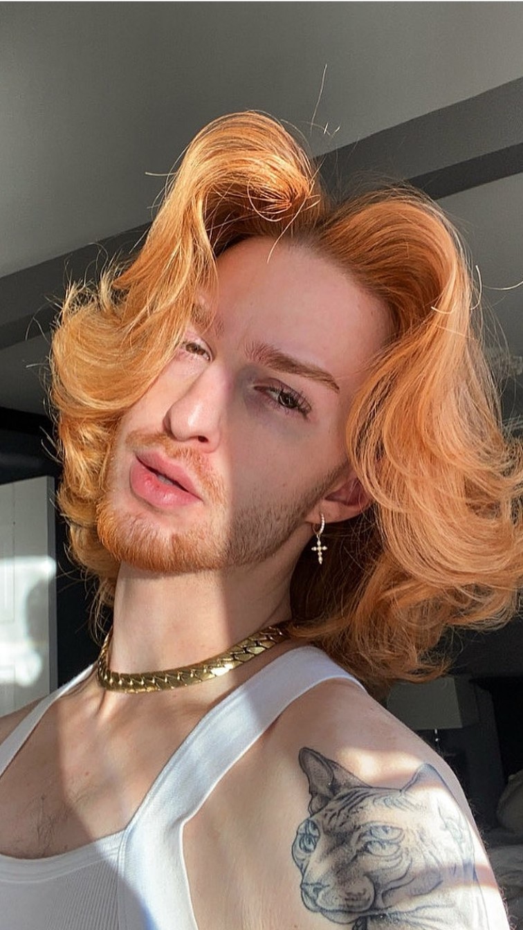 Image of non-binary person with short, wavy ginger hair, a beard, and a thick gold necklace.
